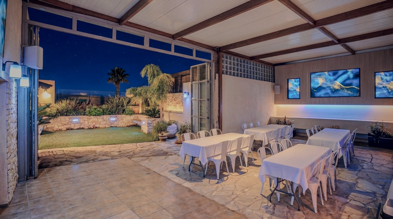 Covered patio with white-tablecloth dining tables overlooking a palm-tree garden at twilight